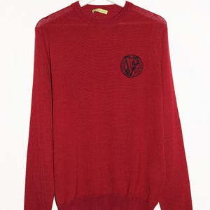 Puede incluir: Un suéter rojo de manga larga con un logotipo Versace en blanco y negro en el pecho izquierdo. El logotipo es un círculo con una cabeza de Medusa dentro.