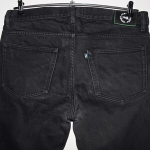 May include: Back view of black denim jeans hanging on a hanger. The jeans have two back pockets and a small logo patch on the waistband. The denim appears to be slightly faded.