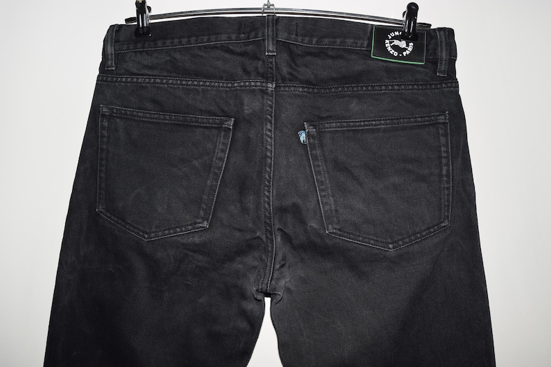 May include: Back view of black denim jeans hanging on a hanger. The jeans have two back pockets and a small logo patch on the waistband. The denim appears to be slightly faded.