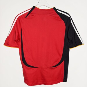 May include: Red soccer jersey with black and white stripes on the sleeves. The jersey has a black and white design on the front and back.