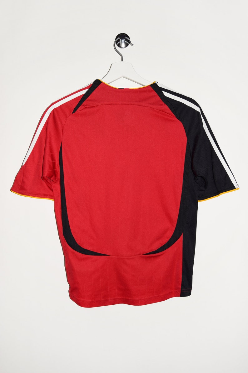 May include: Red soccer jersey with black and white stripes on the sleeves. The jersey has a black and white design on the front and back.