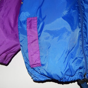 May include: A blue and purple nylon jacket with a zip closure and a drawstring at the bottom. The jacket has a purple pocket on the right side.
