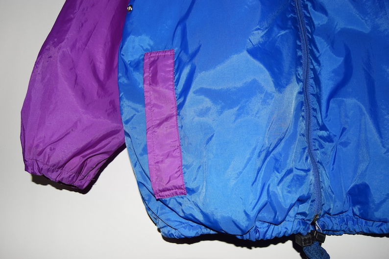 May include: A blue and purple nylon jacket with a zip closure and a drawstring at the bottom. The jacket has a purple pocket on the right side.