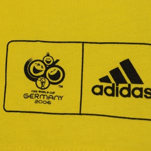 May include: Yellow t-shirt with a black and white logo for the 2006 FIFA World Cup in Germany. The logo features three smiling faces and a soccer ball. The Adidas logo is also on the shirt.