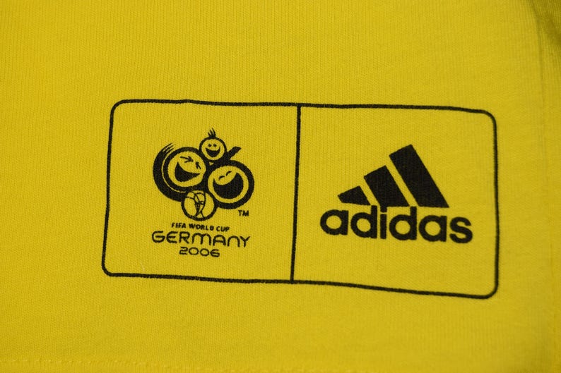 May include: Yellow t-shirt with a black and white logo for the 2006 FIFA World Cup in Germany. The logo features three smiling faces and a soccer ball. The Adidas logo is also on the shirt.