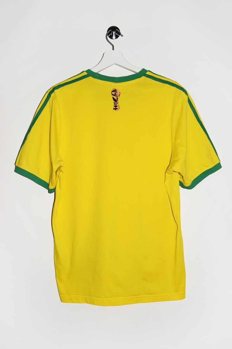 May include: Yellow short-sleeved t-shirt with green trim around the collar and sleeves. The shirt has a small graphic of a soccer trophy on the back.