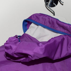 May include: Purple hooded jacket with a blue zipper and a white lining. The jacket is made of a lightweight, water-resistant material.