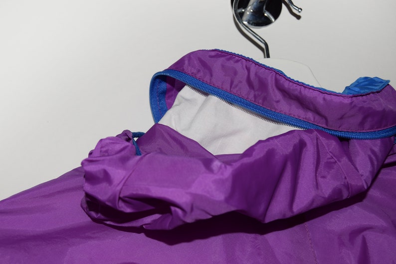 May include: Purple hooded jacket with a blue zipper and a white lining. The jacket is made of a lightweight, water-resistant material.