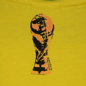 May include: A yellow t-shirt with a black and orange silhouette of the FIFA World Cup trophy. The text "FIFA TM" is printed on the trophy.