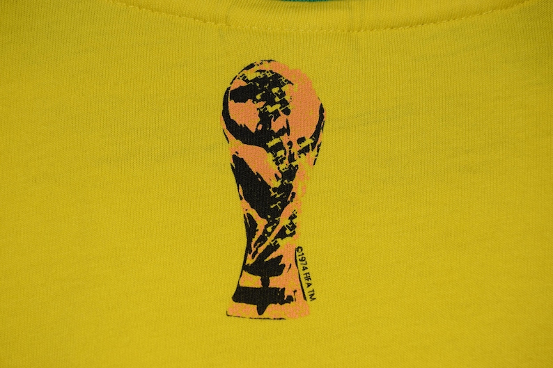 May include: A yellow t-shirt with a black and orange silhouette of the FIFA World Cup trophy. The text "FIFA TM" is printed on the trophy.