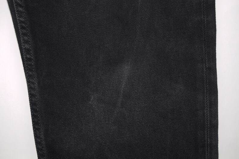 May include: Close-up of black denim fabric. The image shows the texture of the denim, with visible diagonal twill lines. The fabric appears to be a solid black color, with subtle variations in tone. The stitching is visible along the edge of the fabric.