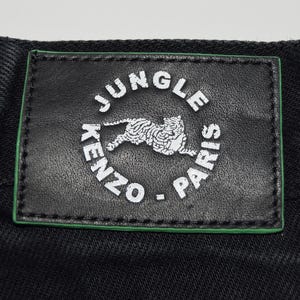 May include: Close-up of black denim fabric with a rectangular black leather patch. The patch features the words "JUNGLE PARIS" and "KENZO" in white, encircling a white tiger illustration, with a green border.