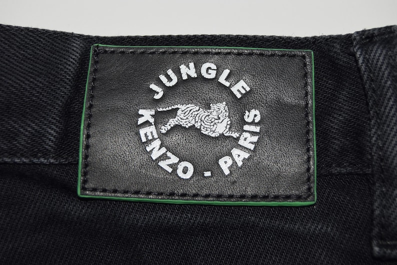 May include: Close-up of black denim fabric with a rectangular black leather patch. The patch features the words "JUNGLE PARIS" and "KENZO" in white, encircling a white tiger illustration, with a green border.