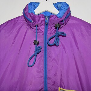 May include: A purple and blue windbreaker jacket with a zip fastening and a drawstring hood. The jacket has a blue lining and a yellow accent on the bottom.