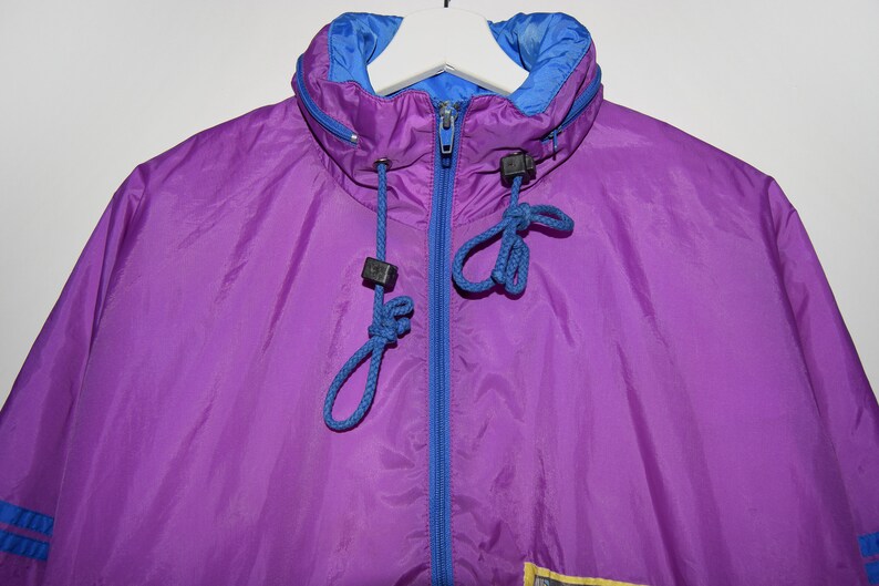 May include: A purple and blue windbreaker jacket with a zip fastening and a drawstring hood. The jacket has a blue lining and a yellow accent on the bottom.