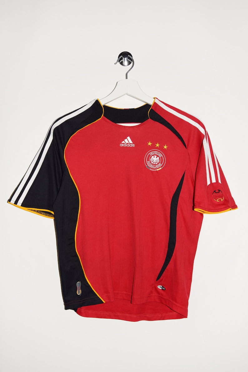 May include: A red soccer jersey with black and white stripes on the sleeves. The jersey has the Adidas logo, three stars, and the German national team crest. The jersey is hanging on a hanger.