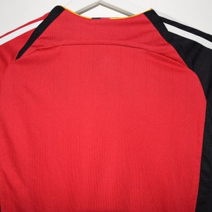 May include: Red soccer jersey with black and white stripes on the sleeves. The jersey has a yellow collar and a black and yellow logo on the back.