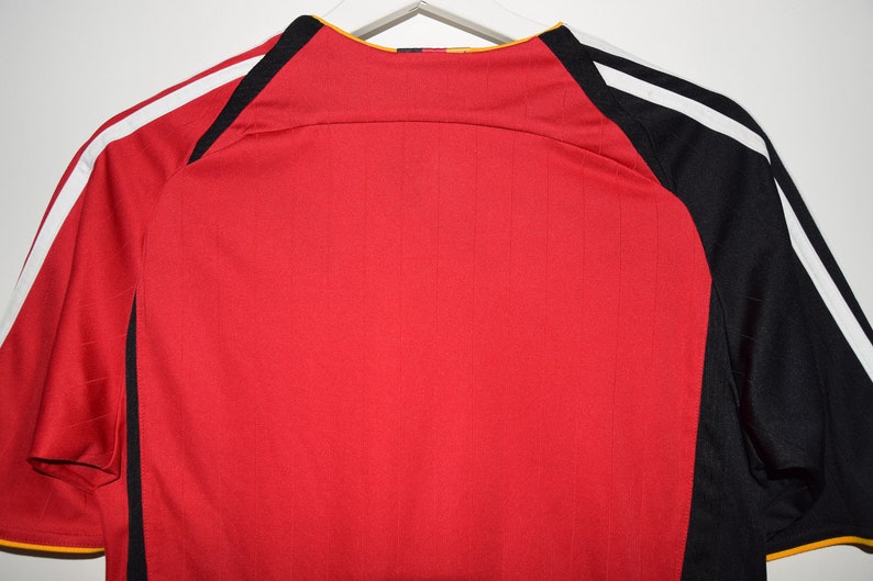 May include: Red soccer jersey with black and white stripes on the sleeves. The jersey has a yellow collar and a black and yellow logo on the back.