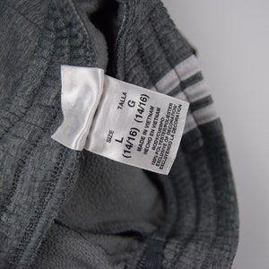 Puede incluir: Una etiqueta blanca con texto negro que indica que la talla es L (14/16) y la talla es G (14/16). La etiqueta tambi&eacute;n dice "Hecho en Vietnam" y "Hecho en Vietnam". La etiqueta est&aacute; unida a un pantal&oacute;n de ch&aacute;ndal gris.