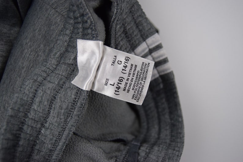 Puede incluir: Una etiqueta blanca con texto negro que indica que la talla es L (14/16) y la talla es G (14/16). La etiqueta tambi&eacute;n dice "Hecho en Vietnam" y "Hecho en Vietnam". La etiqueta est&aacute; unida a un pantal&oacute;n de ch&aacute;ndal gris.