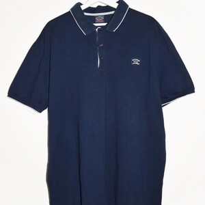 May include: Navy blue polo shirt with white trim on the collar and sleeves. The shirt has a three-button placket and a small logo on the chest. The shirt is hanging on a white hanger.
