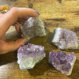 May include: Four amethyst crystal clusters in varying shades of purple and white are displayed on a wooden surface. One cluster is being held by a hand. The crystals have a natural, rough texture.