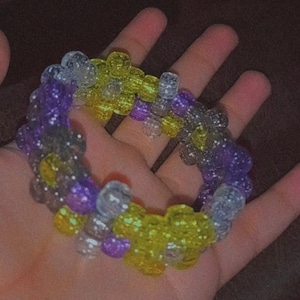 May include: A multi-colored beaded bracelet with purple, yellow, and gray beads. The beads are in a flower shape and have a glittery finish.