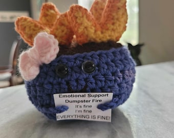 May include: A crocheted plush toy shaped like a head with a blue body, orange hair, and a pink bow. The toy has googly eyes and a sign that reads "Emotional Support Dumpster Fire It's fine I'm fine EVERYTHING IS FINE!"