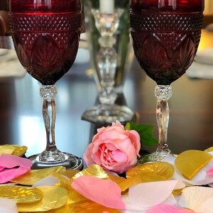 May include: Two ruby red wine glasses with diamond-cut patterns and crystal stems. A pink rose and scattered gold, pink, and white petals are in the foreground. A crystal candle holder and a white candle are in the background.