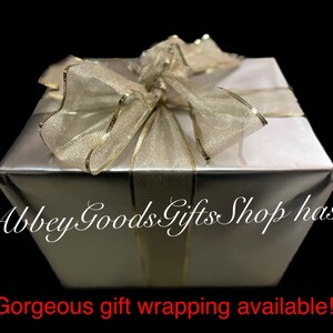 May include: A silver gift box with a gold ribbon and bow. The box is wrapped in shiny silver paper and tied with a sheer gold ribbon. The text "Abbey Goods Gifts Shop has... Gorgeous gift wrapping available!" is visible.