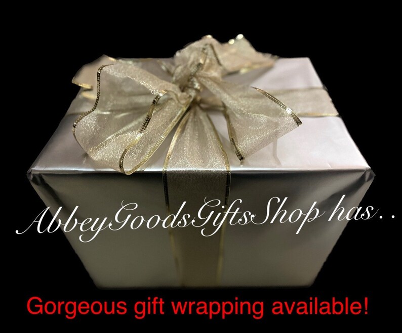 May include: A silver gift box with a gold ribbon and bow. The box is wrapped in shiny silver paper and tied with a sheer gold ribbon. The text "Abbey Goods Gifts Shop has... Gorgeous gift wrapping available!" is visible.