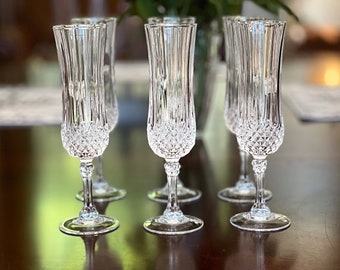 May include: Six clear crystal champagne flutes with a diamond-cut pattern on the lower half and vertical lines on the upper half. The flutes have a long, slender stem and a round base, perfect for celebratory occasions.