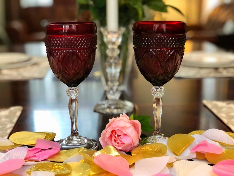 May include: Two ruby red wine glasses with intricate cut glass designs. The glasses sit on a table with a pink rose and scattered gold, pink, and white petals. A crystal candle holder and a lit candle are in the background.