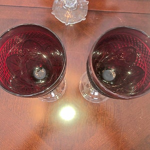 May include: Two ruby red wine glasses with intricate cut glass patterns. The glasses have a classic design with a round bowl and a stemmed base. The glasses are placed on a dark wooden surface, with a decorative placemat visible on the side.