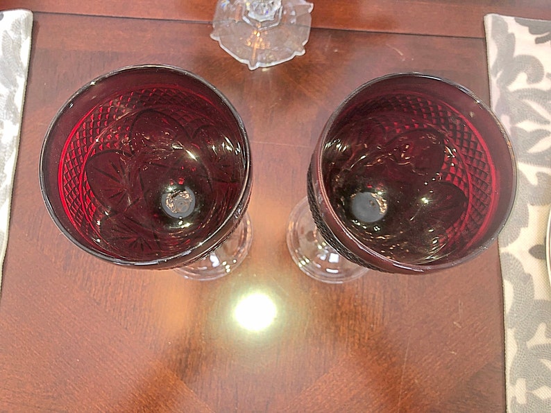 May include: Two ruby red wine glasses with intricate cut glass patterns. The glasses have a classic design with a round bowl and a stemmed base. The glasses are placed on a dark wooden surface, with a decorative placemat visible on the side.