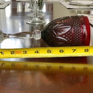 May include: A ruby red wine glass with a clear stem and base, measuring approximately 8 inches in length. The glass features a cut pattern design on the bowl and is positioned horizontally on a wooden surface.