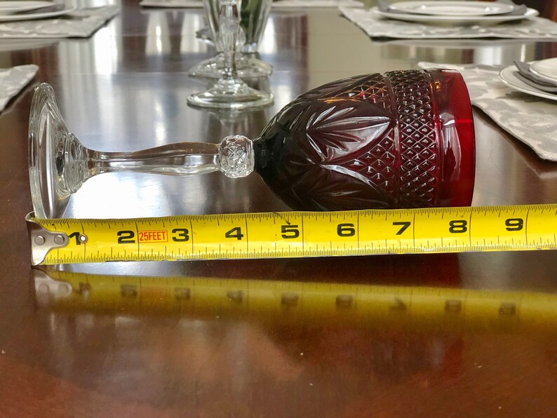 May include: A ruby red wine glass with a clear stem and base, measuring approximately 8 inches in length. The glass features a cut pattern design on the bowl and is positioned horizontally on a wooden surface.