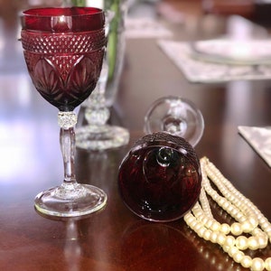 May include: A ruby red wine glass with intricate cut glass detailing, a clear stem, and a decorative base. A second glass lies on its side. A strand of faux pearls rests nearby, suggesting a vintage or elegant theme.