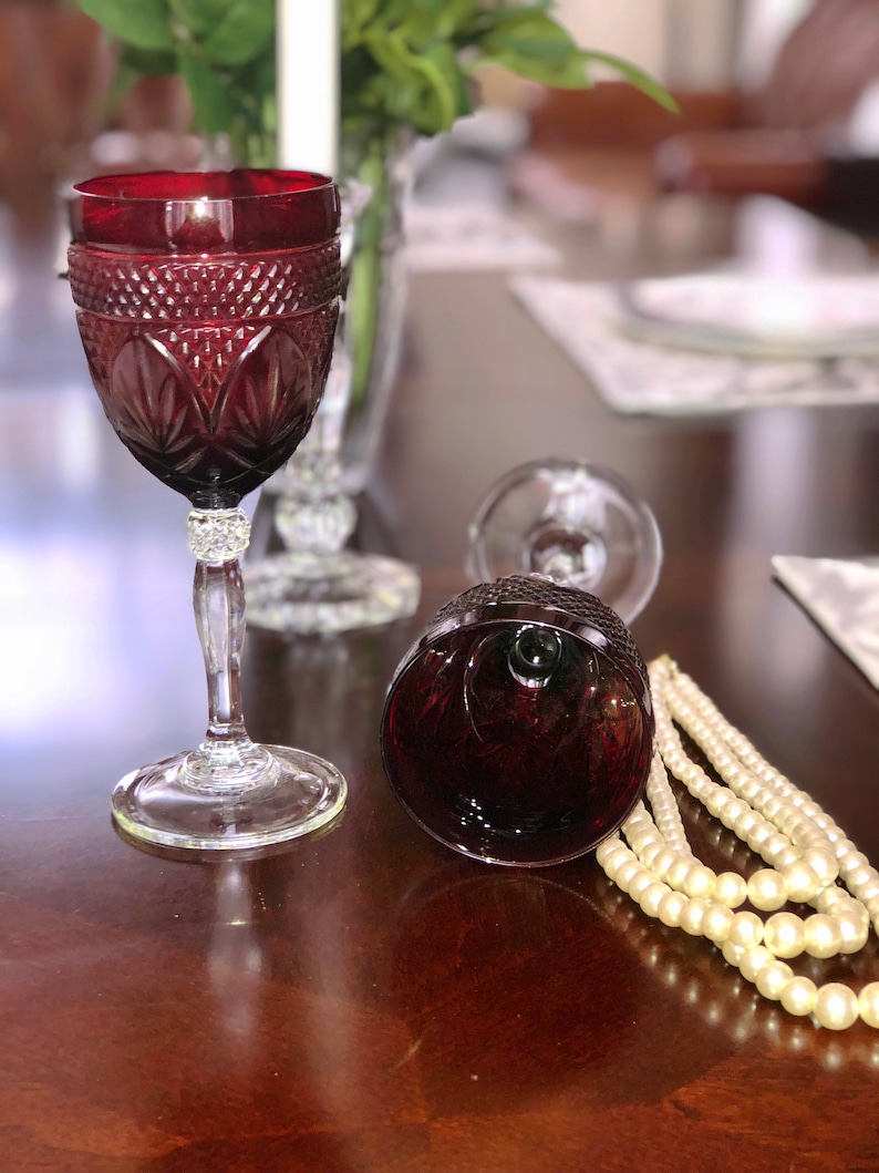 May include: A ruby red wine glass with intricate cut glass detailing, a clear stem, and a decorative base. A second glass lies on its side. A strand of faux pearls rests nearby, suggesting a vintage or elegant theme.