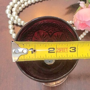 May include: A ruby red glass with a cut pattern, measuring approximately 2.5 inches in diameter. The glass has a decorative design and sits on a clear base. A yellow measuring tape is placed across the top of the glass.