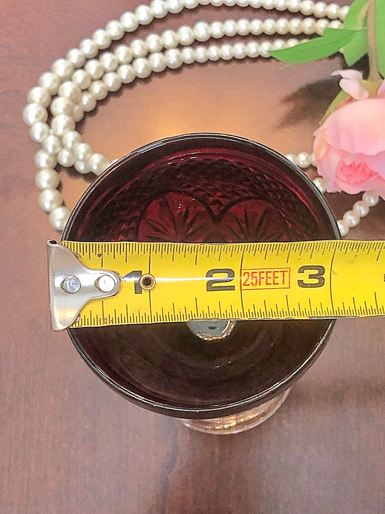 May include: A ruby red glass with a cut pattern, measuring approximately 2.5 inches in diameter. The glass has a decorative design and sits on a clear base. A yellow measuring tape is placed across the top of the glass.