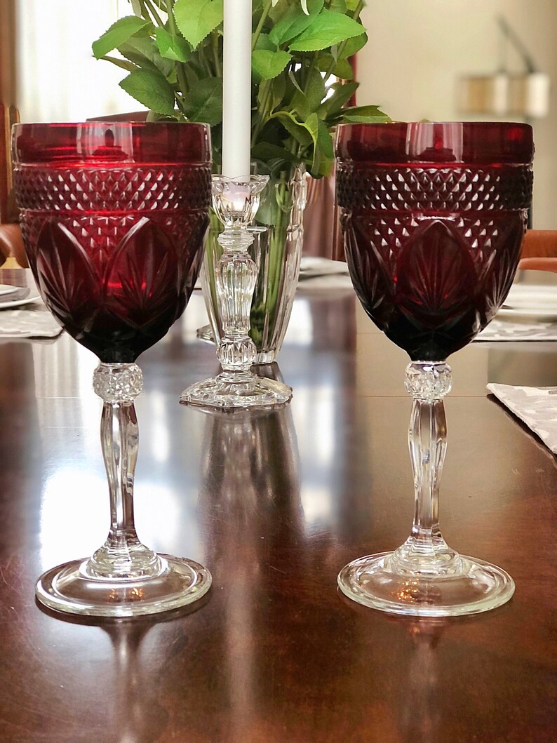 May include: Two ruby red wine glasses with intricate cut glass patterns. The glasses have clear, faceted stems and bases. The image is of a dining table setting, with a candle holder and greenery in the background.