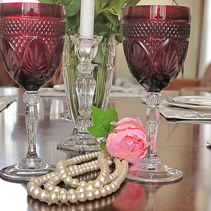 May include: Two ruby red wine glasses with intricate cut glass designs. The glasses sit on a dark wooden table, next to a pearl necklace, a pink rose, and a crystal candlestick holder. The stemware is elegant and decorative.