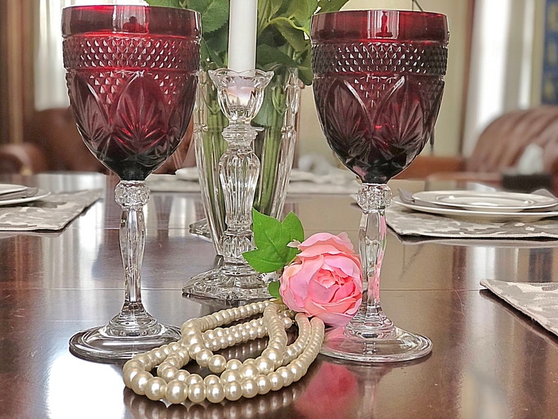 May include: Two ruby red wine glasses with intricate cut glass designs. The glasses sit on a dark wooden table, next to a pearl necklace, a pink rose, and a crystal candlestick holder. The stemware is elegant and decorative.