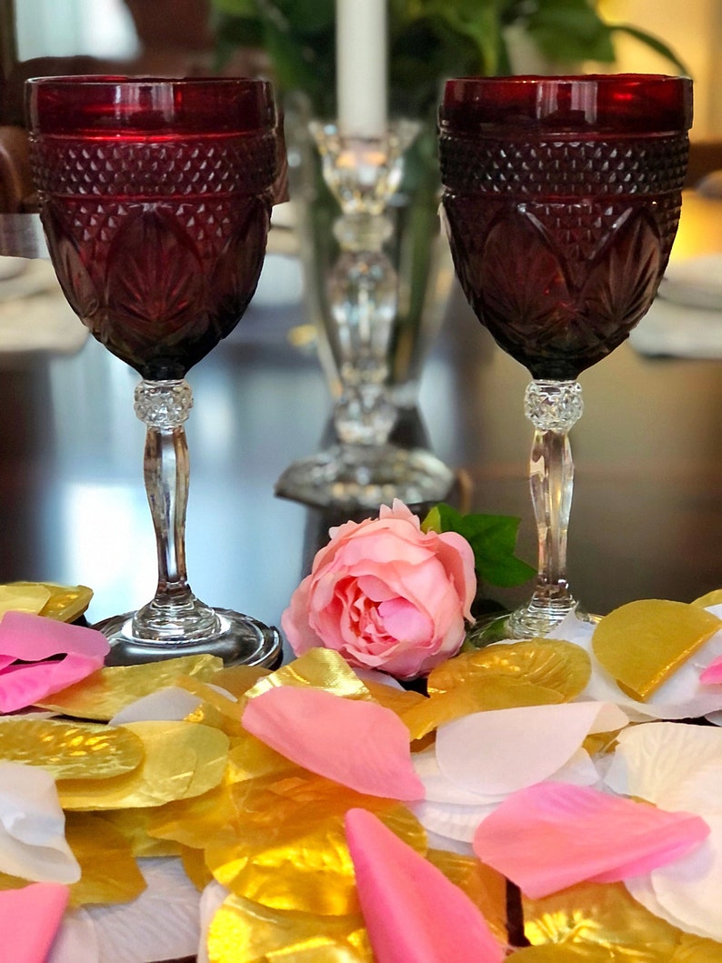 May include: Two ruby red wine glasses with diamond-cut patterns and crystal stems. A pink rose and scattered gold, pink, and white petals are in the foreground. A crystal candle holder and a white candle are in the background.