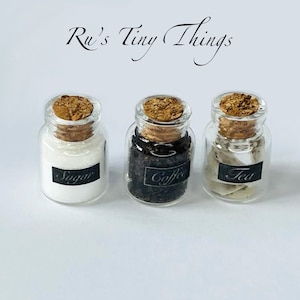May include: Three small glass jars with cork lids and labels. The jars are filled with white sugar, black coffee beans, and white tea leaves. The labels read "Sugar", "Coffee", and "Tea".