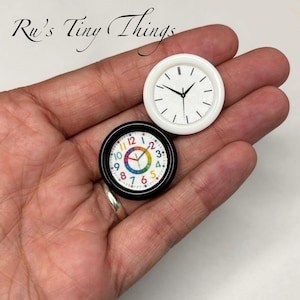 Könnte beinhalten: Zwei Miniaturuhren, eine weiße mit schwarzen Zeigern und die andere schwarz mit einem bunten Regenbogen-Zifferblatt. Die Uhren sind klein, rund und scheinen dekorative Gegenstände zu sein. Der Text "Ru's Tiny Things" befindet sich in der oberen linken Ecke.