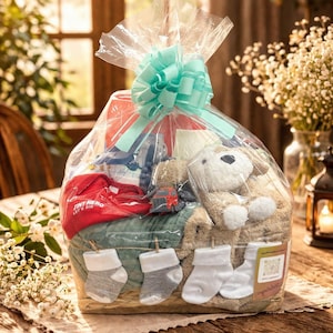 Puede incluir: Una canasta de regalo para bebé envuelta en celofán transparente y atada con un lazo azul claro. La canasta contiene un body rojo con el texto "Tiny Hero", un peluche, calcetines y otros artículos para bebé.