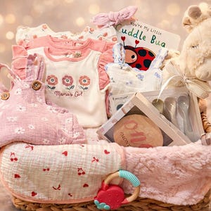 Puede incluir: Una canasta de regalo para bebés llena de ropa rosa y blanca, una jirafa de peluche, un libro y un mordedor de madera. La canasta incluye un peto con estampado floral rosa, una camisa blanca con bordado floral y una manta suave.