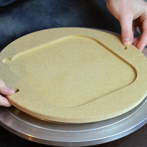 May include: A light brown, circular wooden piece with a square-shaped cutout in the center. The piece has a rounded edge and two small holes. The object is being held by two hands, and it is placed on a silver surface.
